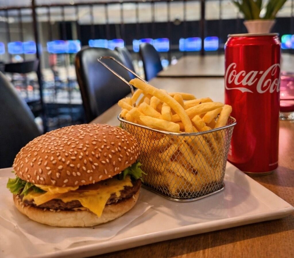 En sesamfrÃ¶hamburgare med ost, sallad och en korg med krispiga pommes frites, tillsammans med en burk Coca-Cola, serverad pÃ¥ ett bord.