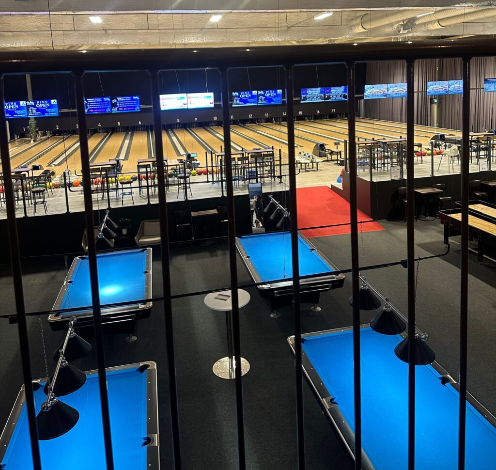 Utsikt Ã¶ver en bowlinghall med flera bowlingbanor och biljardbord i fÃ¶rgrunden. SkÃ¤rmar visar information om event. 